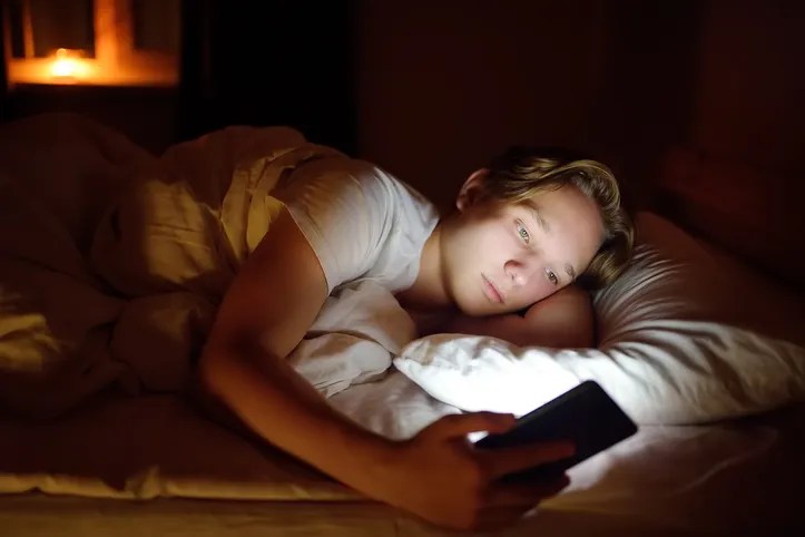 Florida student chatting by phone, writing in blog or reading social networks instead sleeping. Young guy laying in bed and watching video clips by smartphone. Overuse and addiction kids from gadgets.